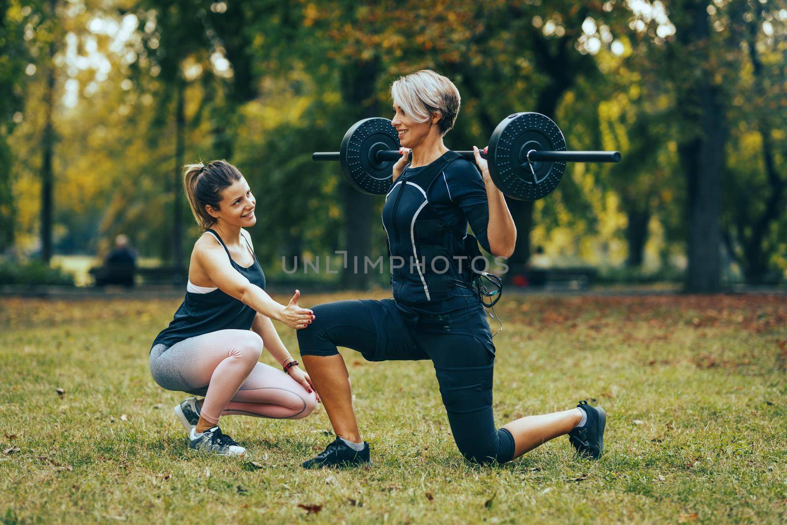 A fit mature woman is doing lunges exercises with personal trainer in the park, dressed in a black suit with an EMS electronic simulator to stimulate her muscles.