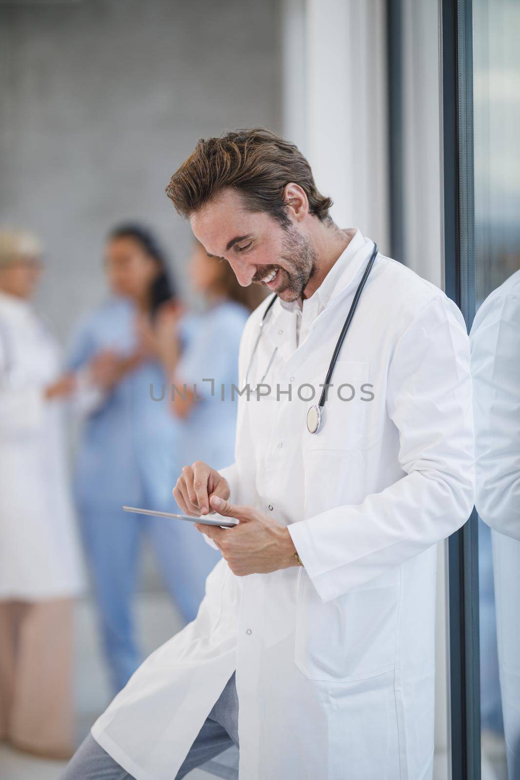 Shot of a smiling doctor standing near a window and using digital tablet while having quick break in a hospital hallway.