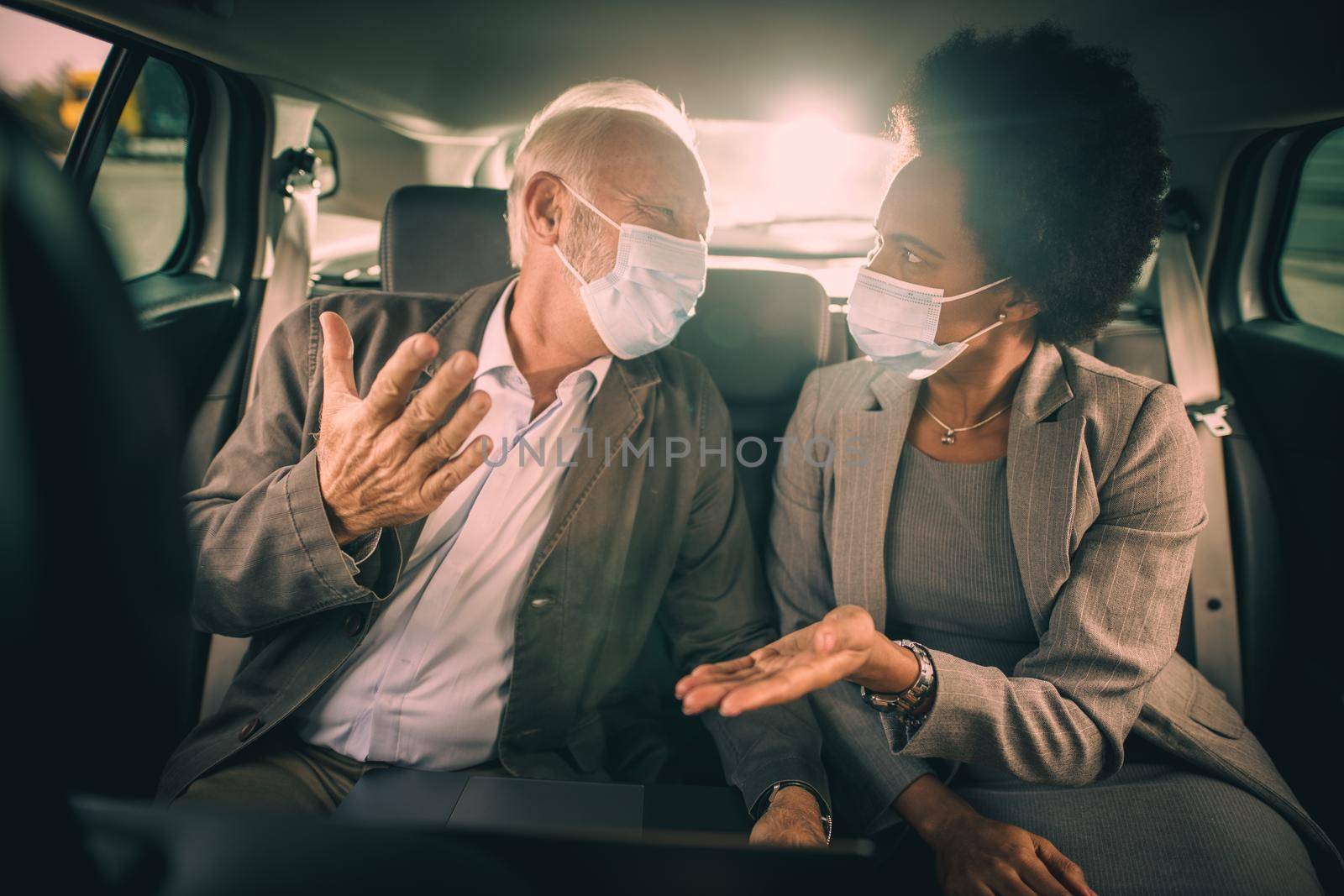 Shot of two successful multi-ethnic people with protective mask using laptop and having a discussion while sitting in the backseat of a car on their business commute during COVID-19 pandemic.