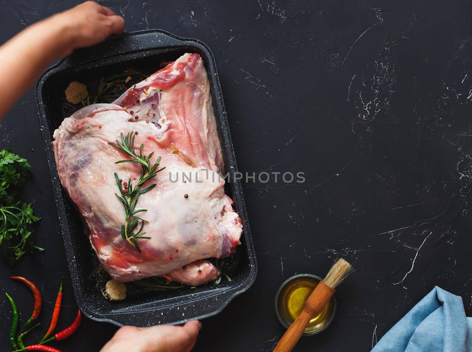 Woman preparing a meat roast with herbs and garlic. Ready to roast shoulder of lamb seasoned with herbs in a roasting tin. Top view, blank space by Slast20
