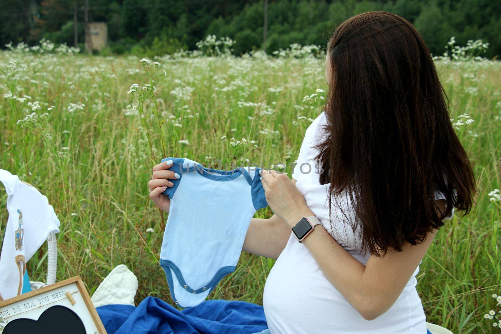 Pregnant woman in white t-shirt holding a blue baby bodysuit in the field