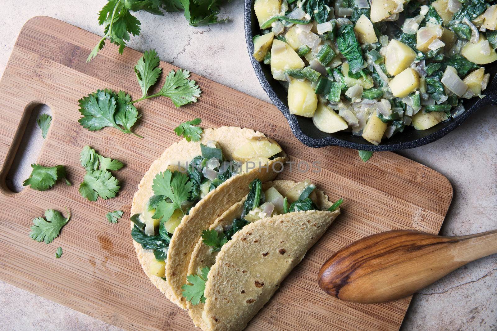 Two vegan soft tacos filled with potatoes and spinach and topped with cilantro on a cutting board with iron pan of filling on the side.