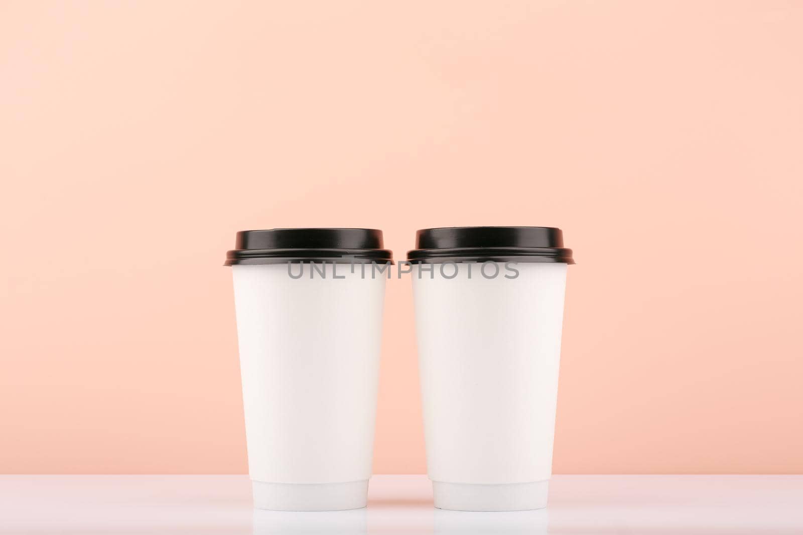 Two white disposable carton coffee or tea cups with black caps on white table against beige background with copy space. Concept of hot drinks