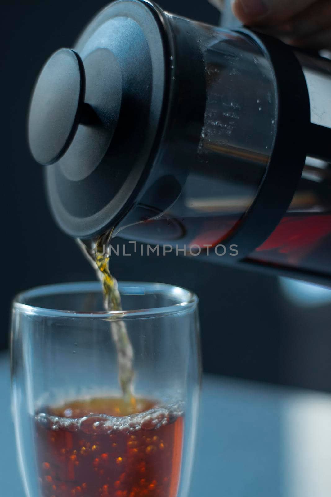 french press black tea pouring into a glass with a double bottom on a white table and grey background - vertical photo