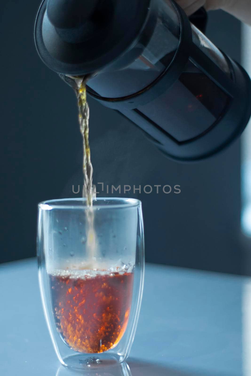 french press black tea pouring into a glass with a double bottom on a white table and grey background - vertical photo