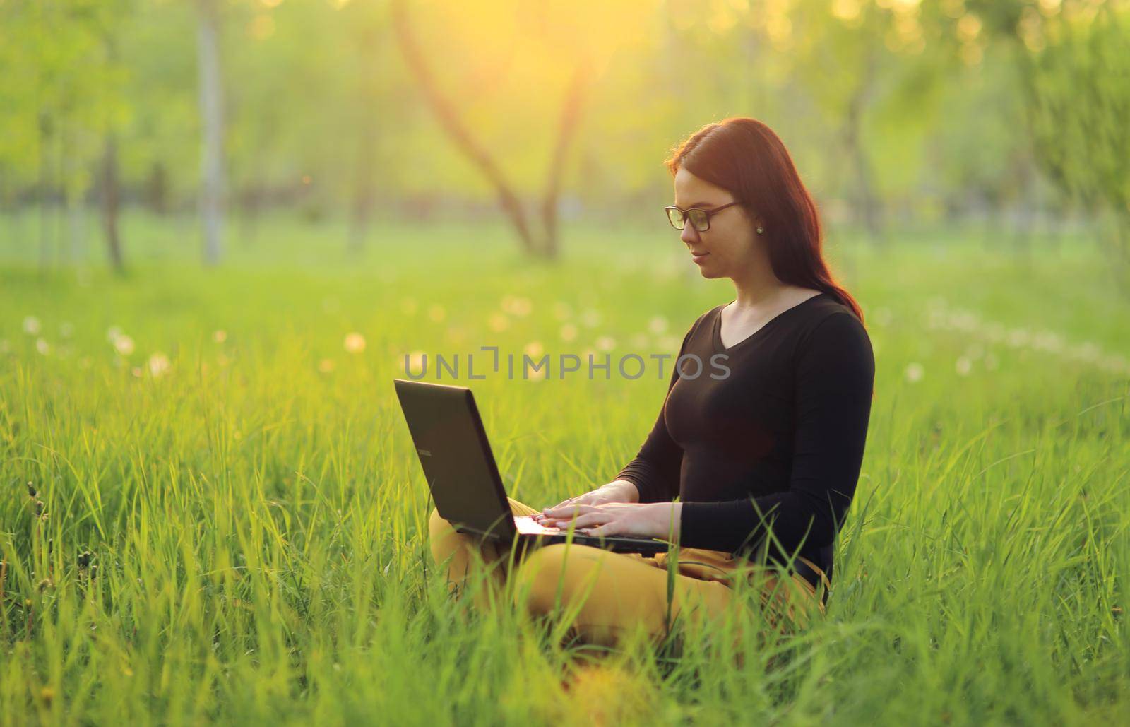 Cute smiling girl with laptop sits on green grass in the park and works remotely as a freelancer by selinsmo
