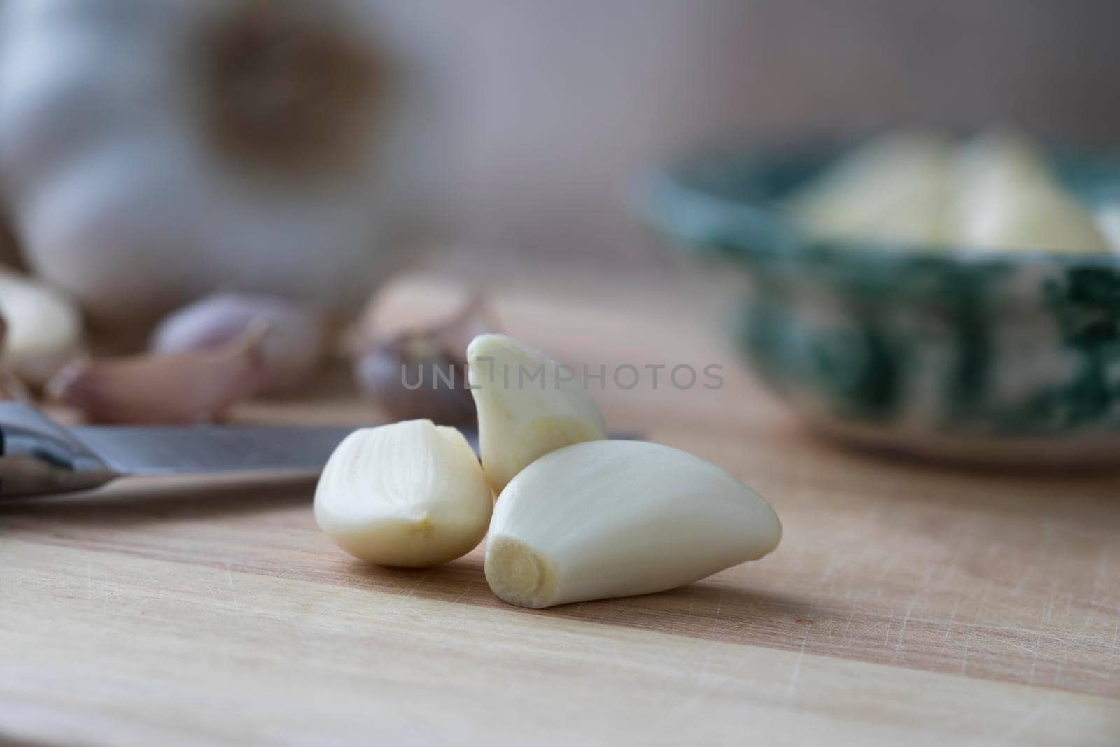Peeled garlic cloves on wooden cutting board with knife and bowl in background.