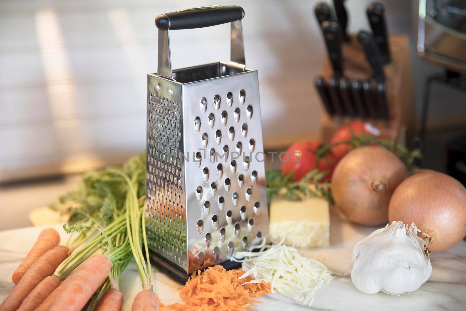 Cheese grater in kitchen surrounded by carrots,  cheese, garlic, onions.