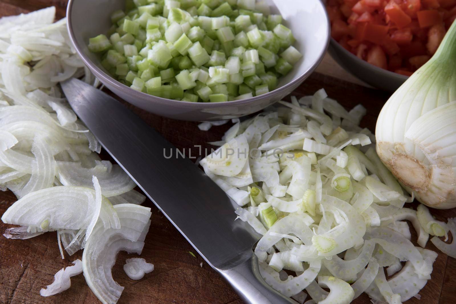 Sliced fennel and yellow onions with bowl of chpped celery and chef's knife on  a cutting board