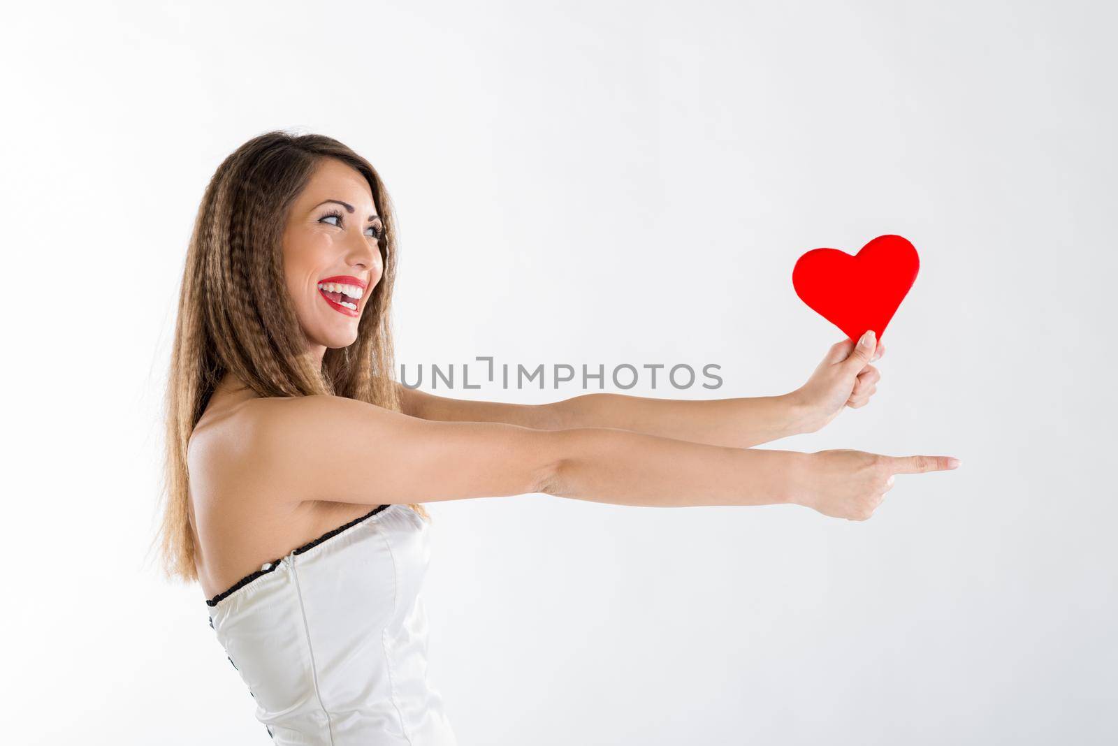Beautiful cheerful girl standing, holding red heart and pointing something.