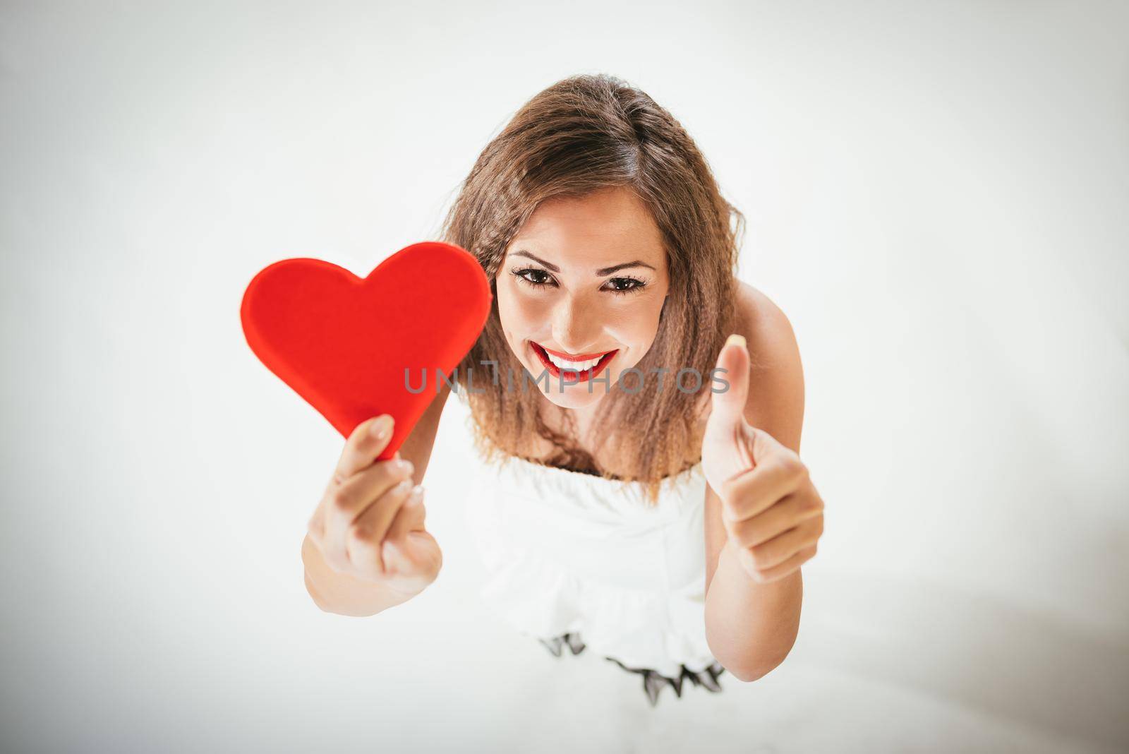 Close-up of a beautiful smiling girl holding a red heart with tumb up. Selective focus.