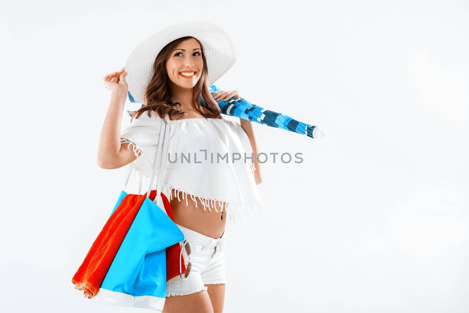 Beautiful young woman on summer vacation. She is standing with hat and holding umbrella and bag for beach. 
