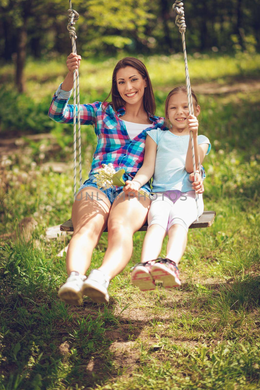 Happy young woman and little girl enjoying on the swing in the spring garden. Looking at camera.