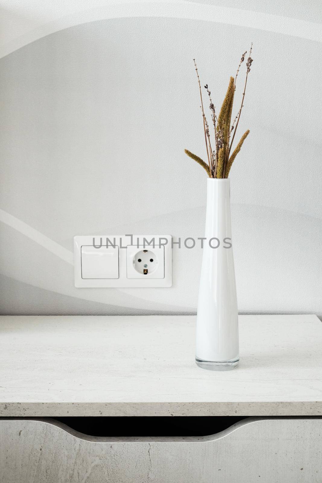 Stylish wooden bedside table and white glass vase with dried branch and plants on it. Interior details. Vertical image.