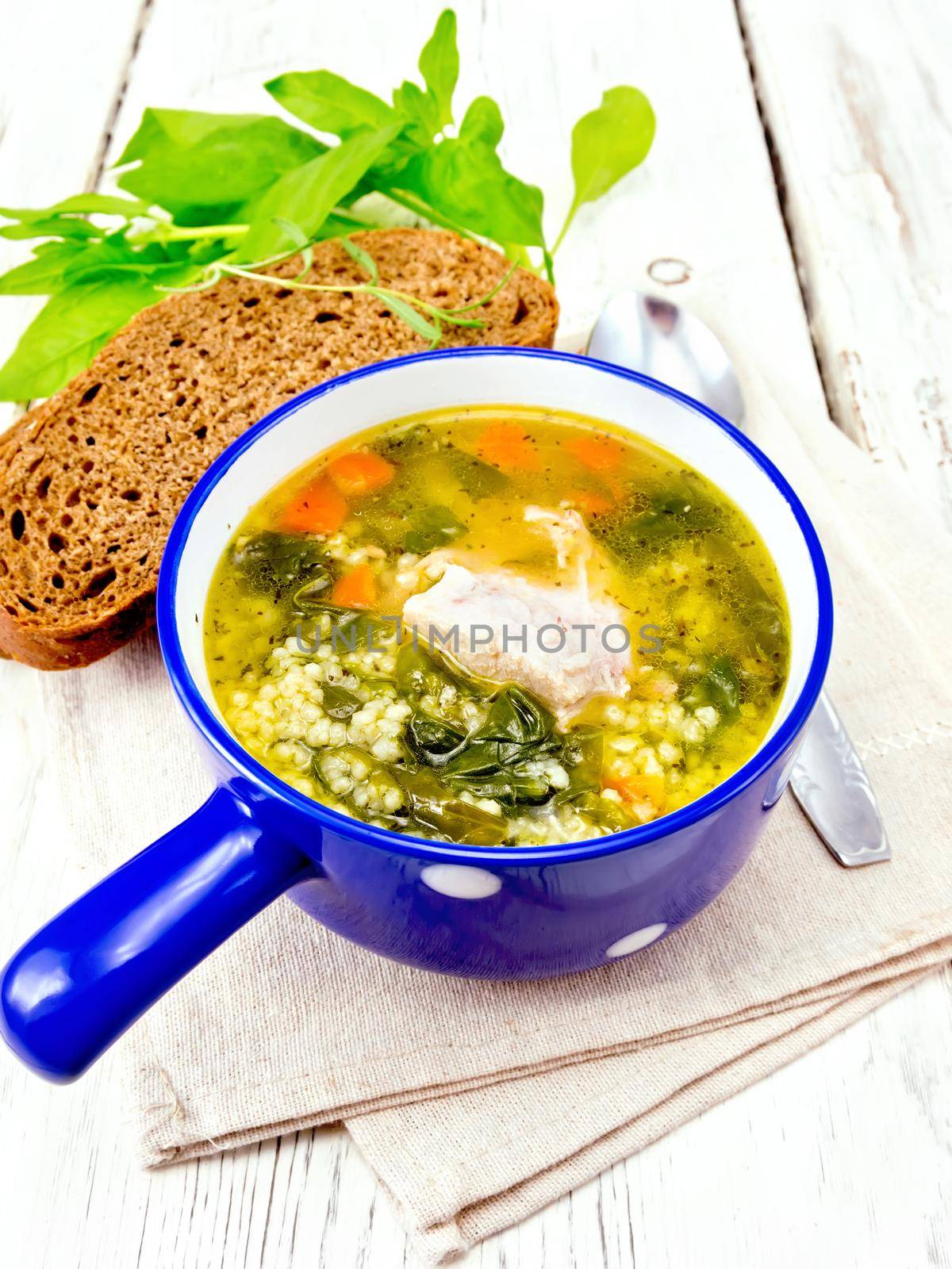 Pork ribs soup with couscous and spinach in a blue bowl on towel, bread on wooden board background