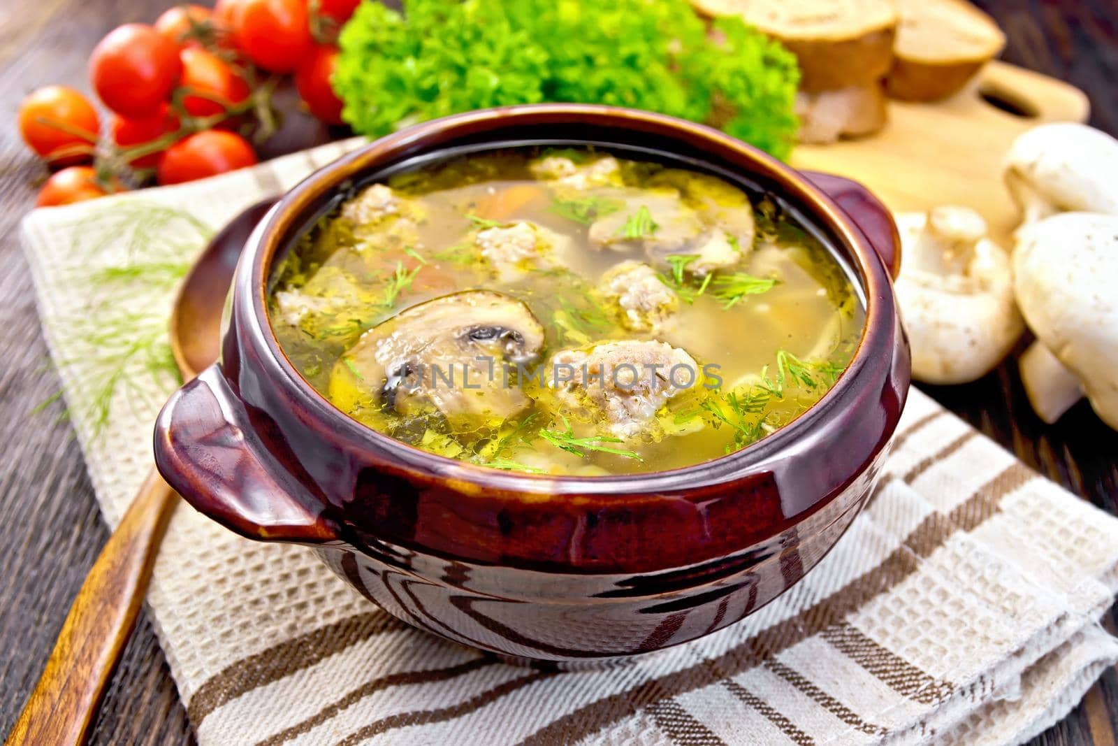 Soup with meatballs, noodles and champignon in a clay bowl on a napkin, parsley, tomatoes, mushrooms and bread on a wooden boards background