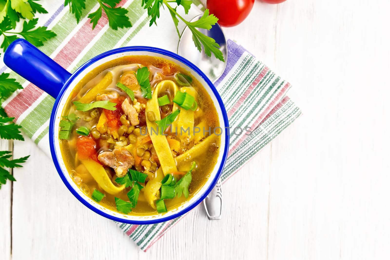 Soup with meat, tomatoes, vegetables, mung bean lentils and noodles in a blue bowl on a towel, parsley and spoon on wooden board background from above