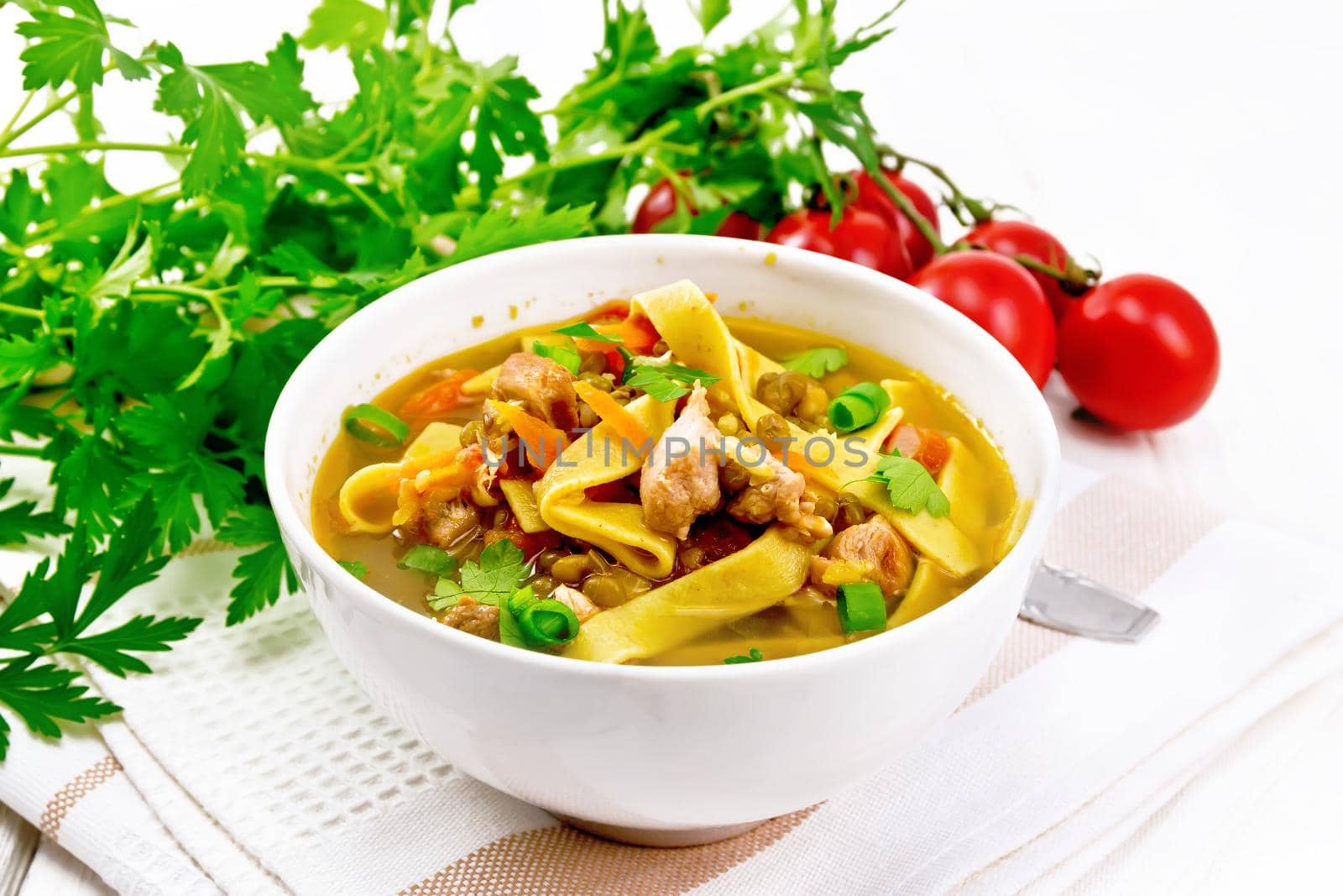 Soup with meat, tomatoes, vegetables, mung bean lentils and noodles in a bowl on a towel, parsley and spoon on wooden board background