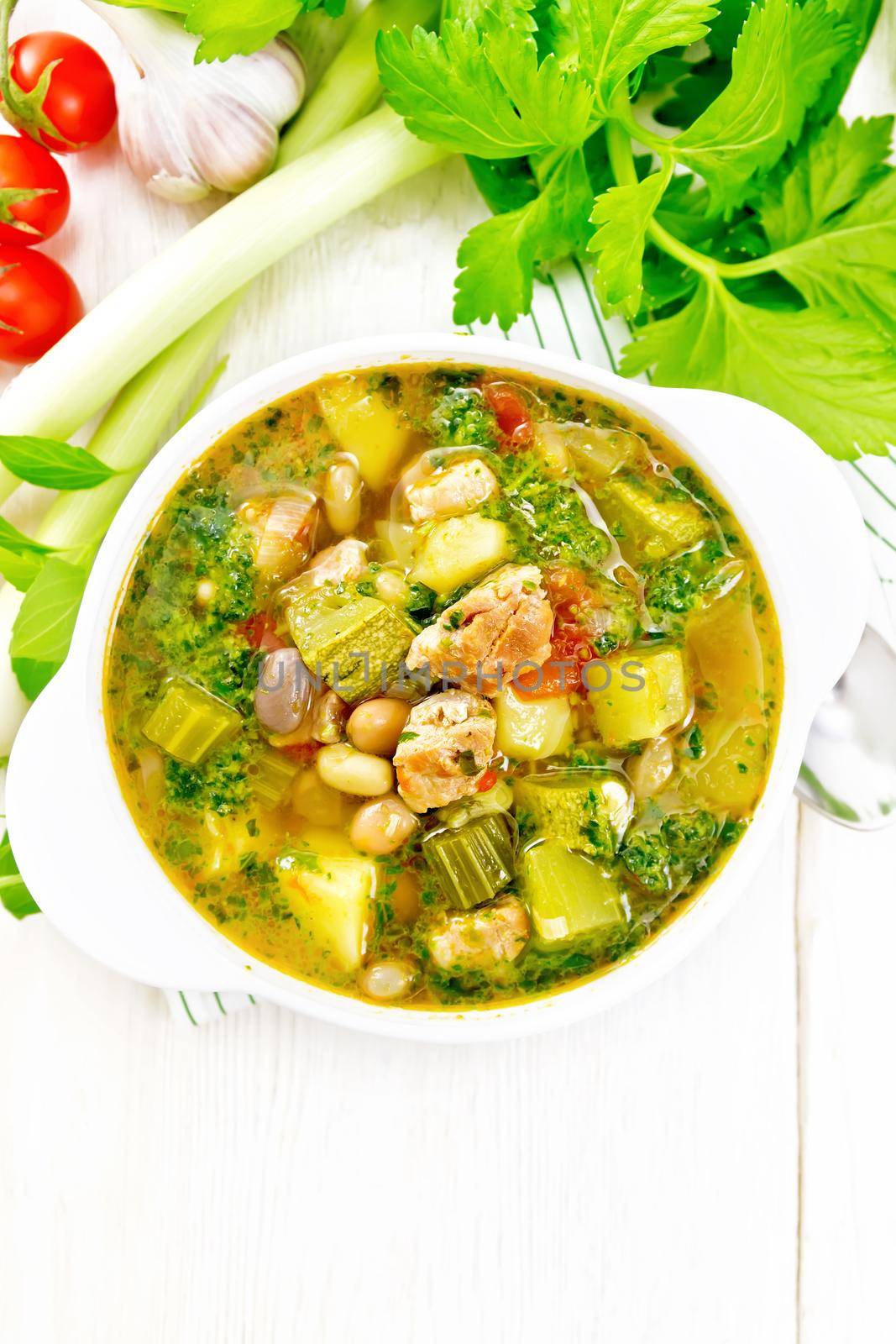 Soup with pisto sauce, lard, tomatoes, zucchini, beans, celery and leeks in a bowl on a towel on wooden board background from above