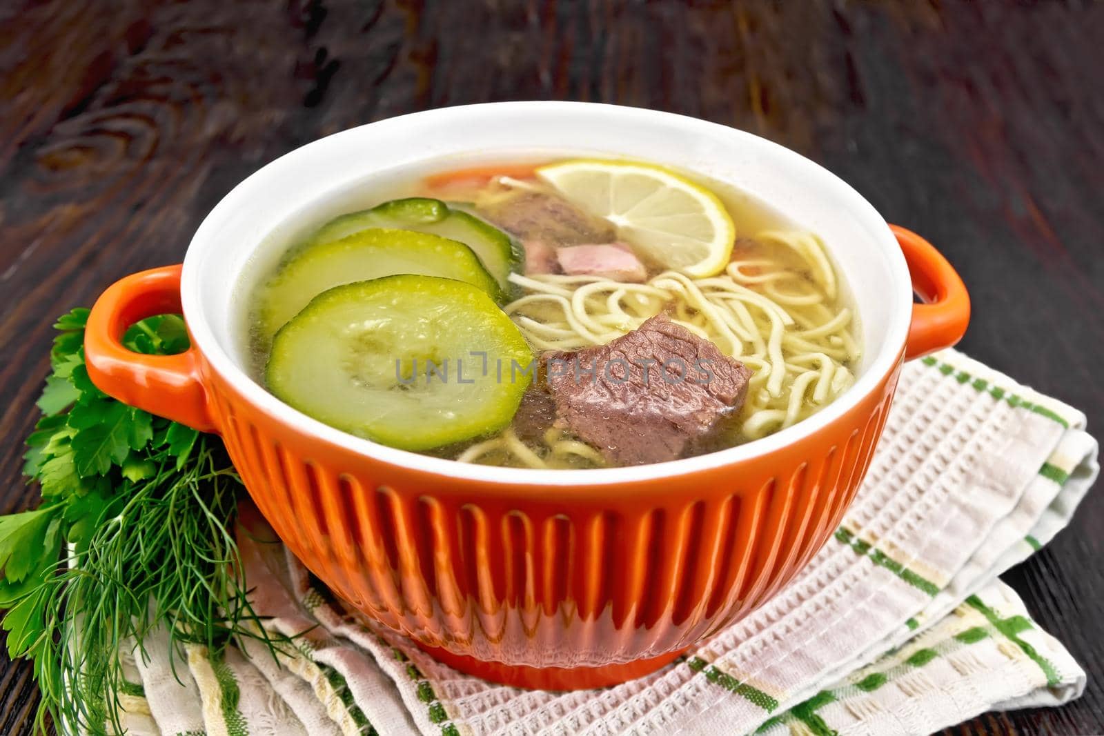 Soup with zucchini, beef, ham, lemon and noodles in a red bowl, parsley and dill on a napkin on a background of a dark wooden board