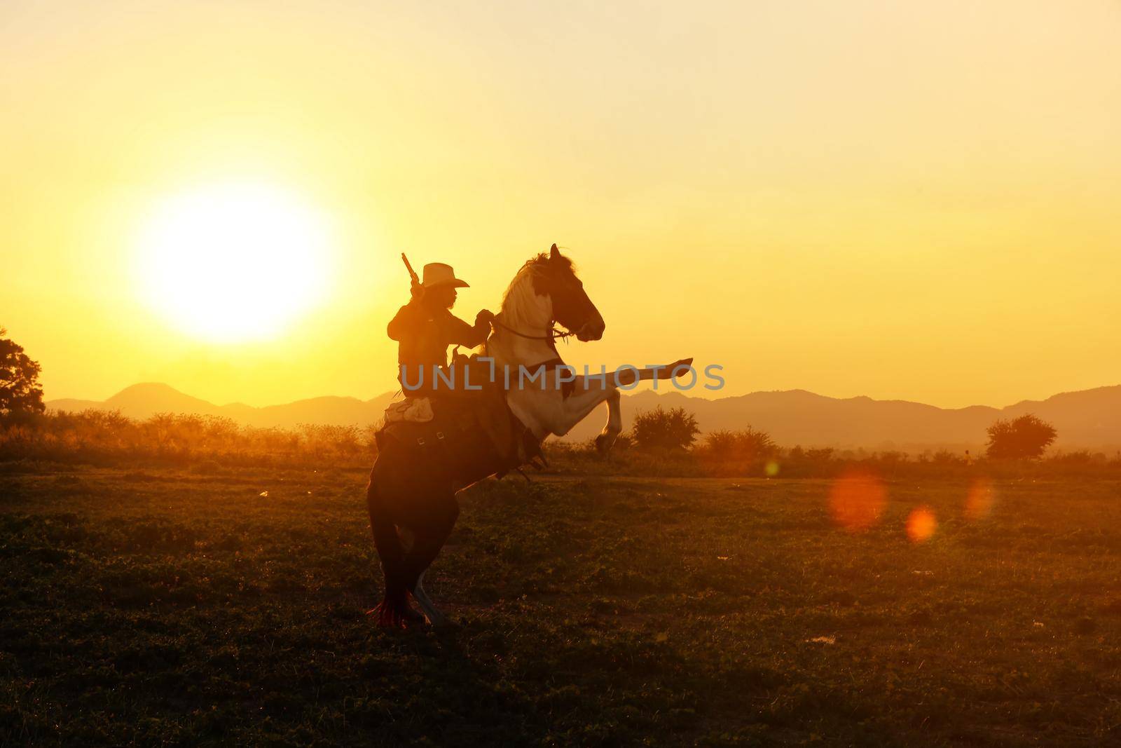 The silhouette of rider as cowboy outfit costume with a horses and a gun held in the hand against smoke and sunset background by chuanchai