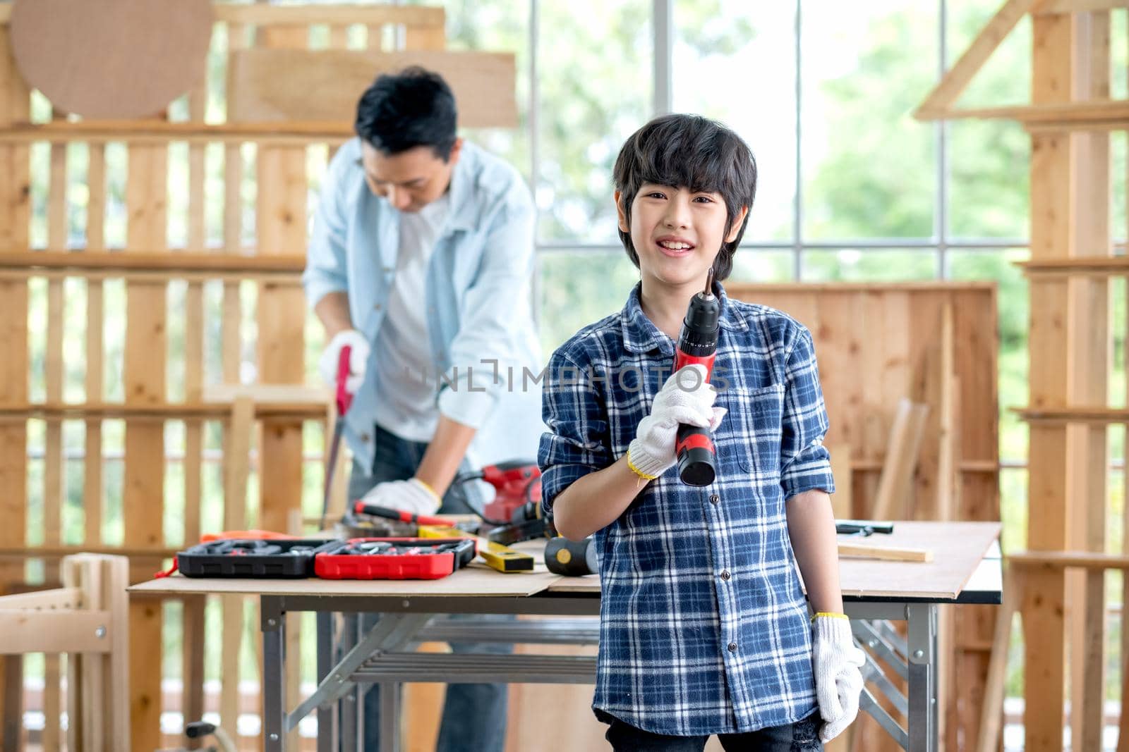 Asian boy hold electric drill and smile with look at camera. His father stay on the background and work as carpentering at home.