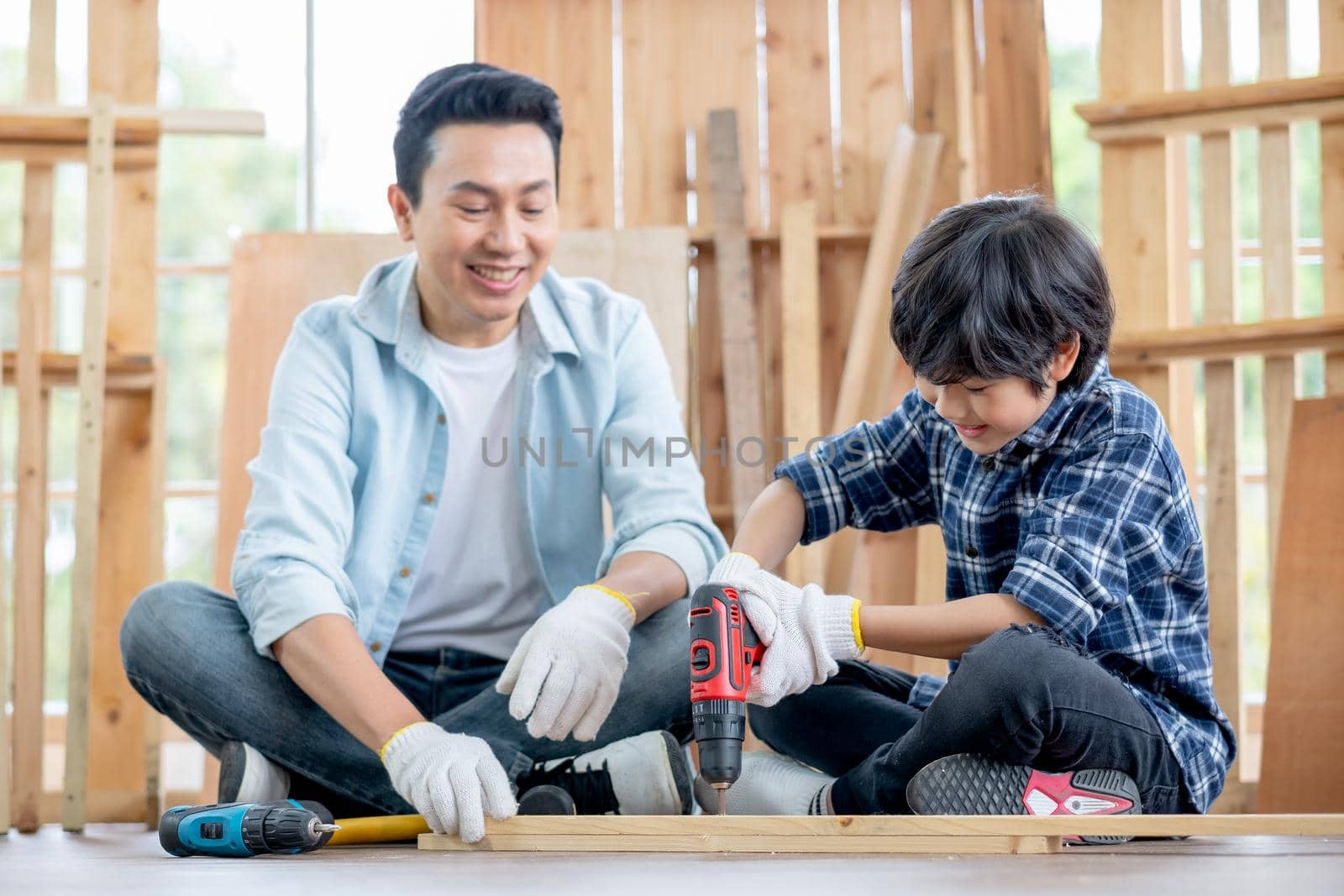Asian father look carefully son use drill in their workplace of carpentering with happy emotion. Asian family concept to stay at home and enjoy good relationship hobby together.