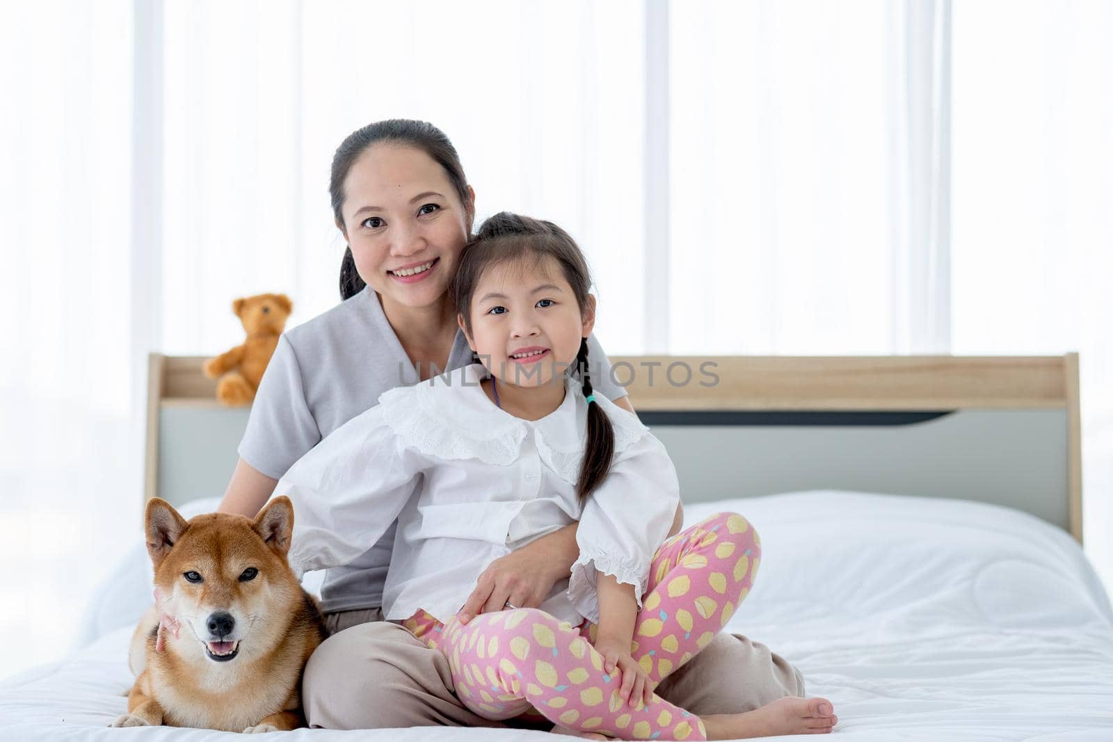 Asian mother and little girl sit on bed with shiba inu dog also look at camera with smiling of happiness.
