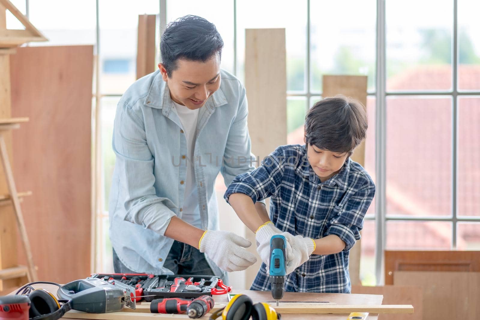 Asian father teach his son to use electric drill to work with woodwork in their house. Concept of good relationship with hobby or activities in happy family.