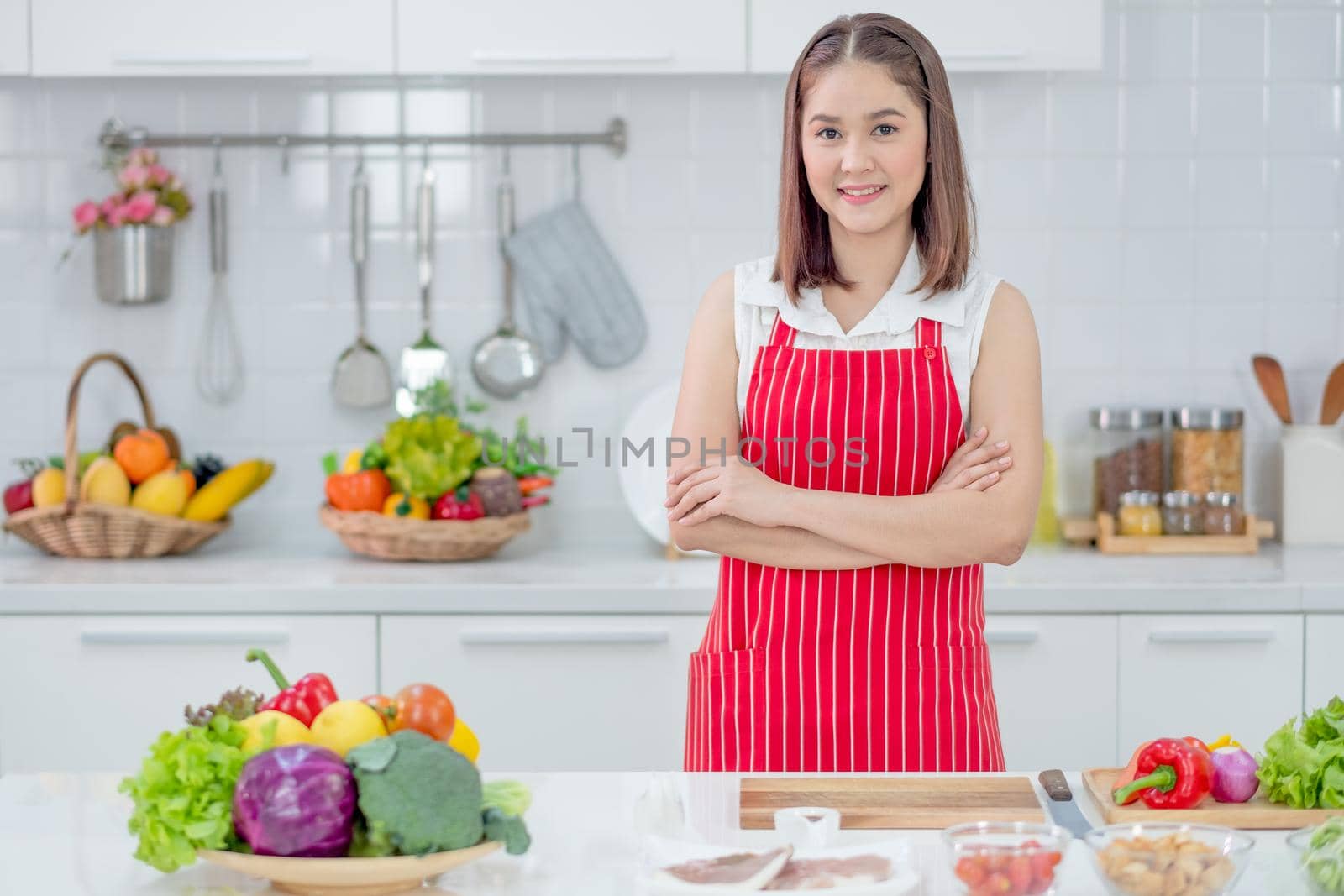 Asian beautiful woman with red apron stand with arm crossed and smiling in kitchen and look to camera with different ingredients and food. Concept of happiness of cooking in their house.