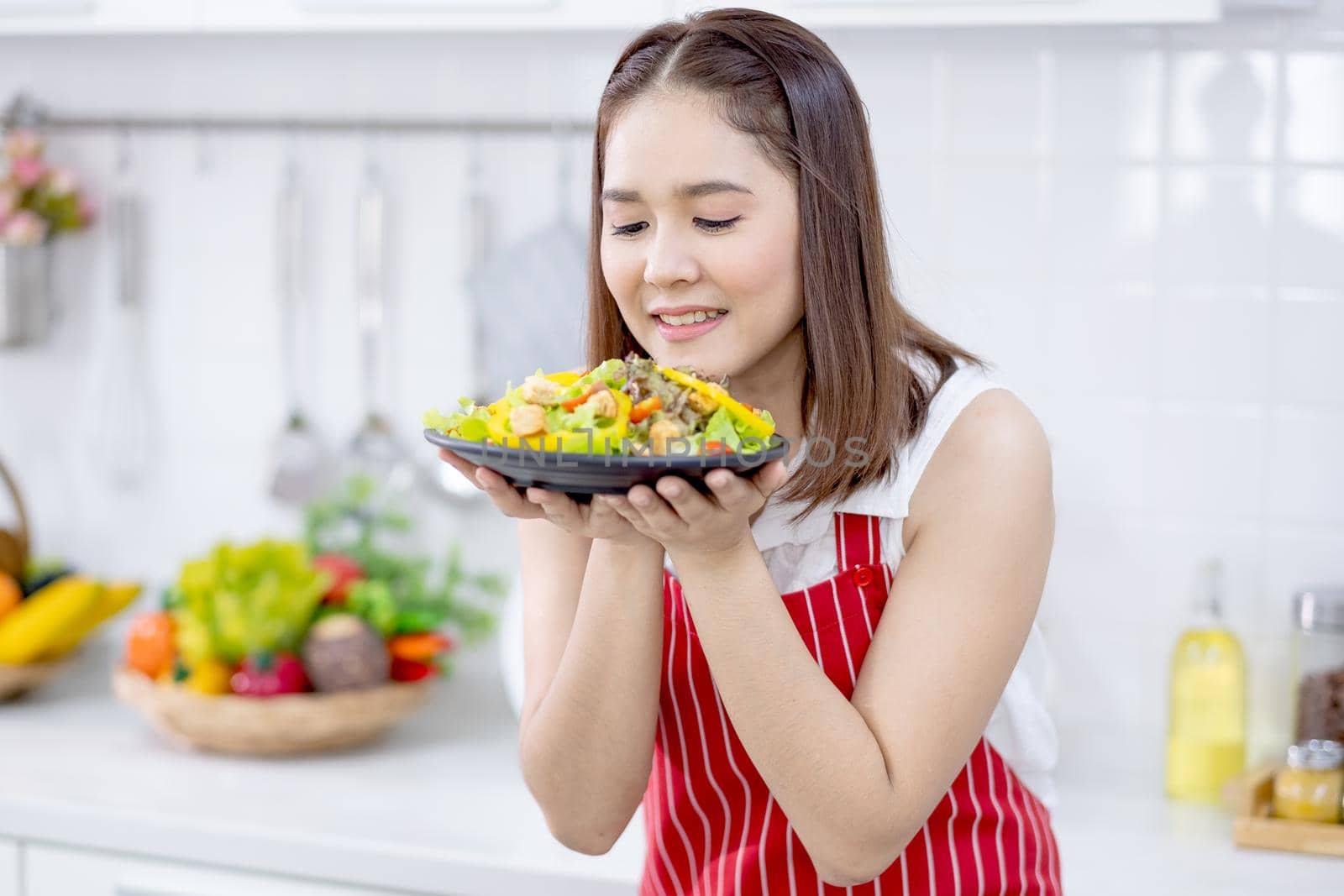 Asian beautiful woman with red apron hold plate with filled in food and action of good smell in kitchen and look to camera with smiling. Concept of happiness of cooking in their house.