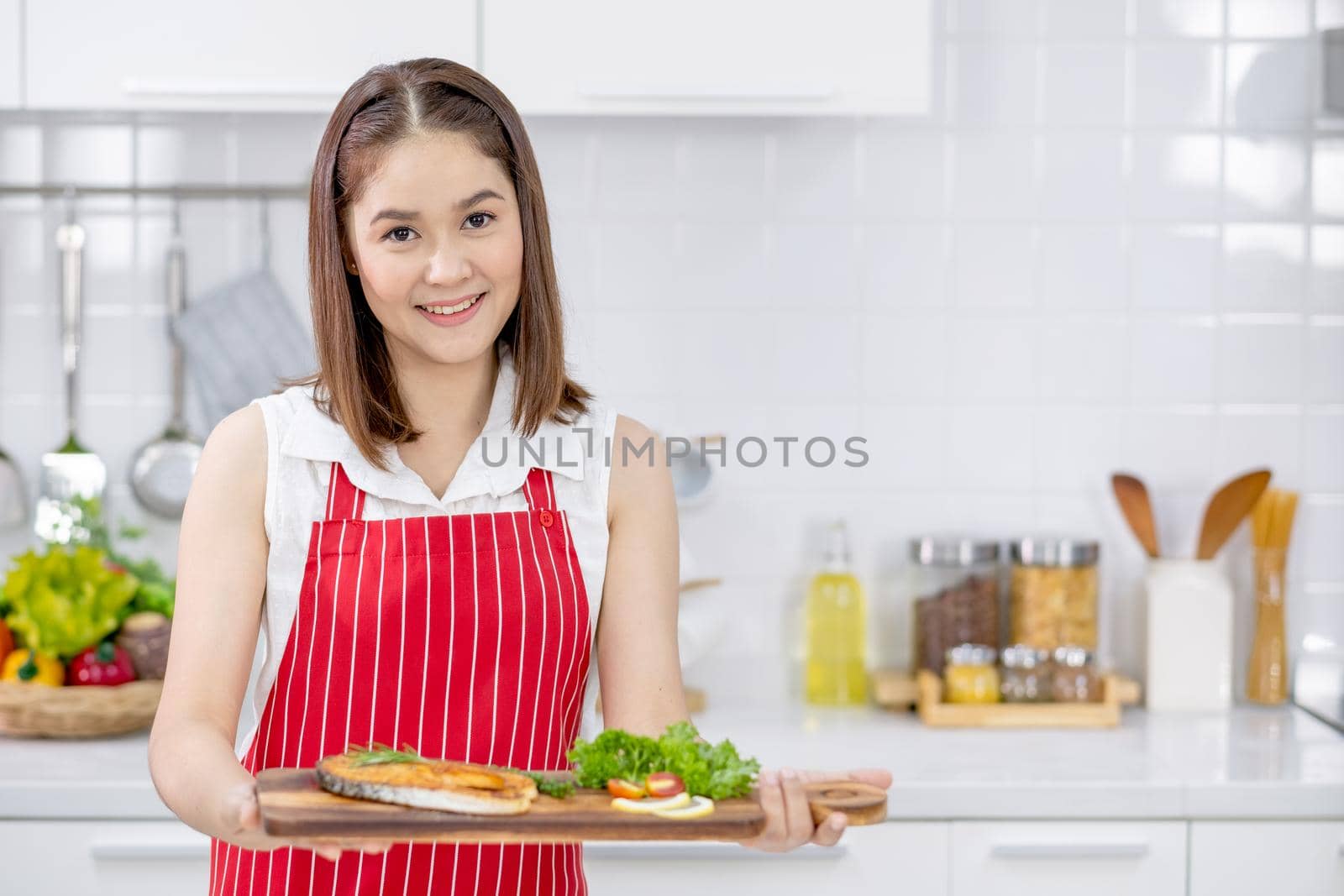 Asian beautiful woman with red apron hold and present slicing fish on plate in kitchen and look to camera with smiling. Concept of happiness of cooking in their house.