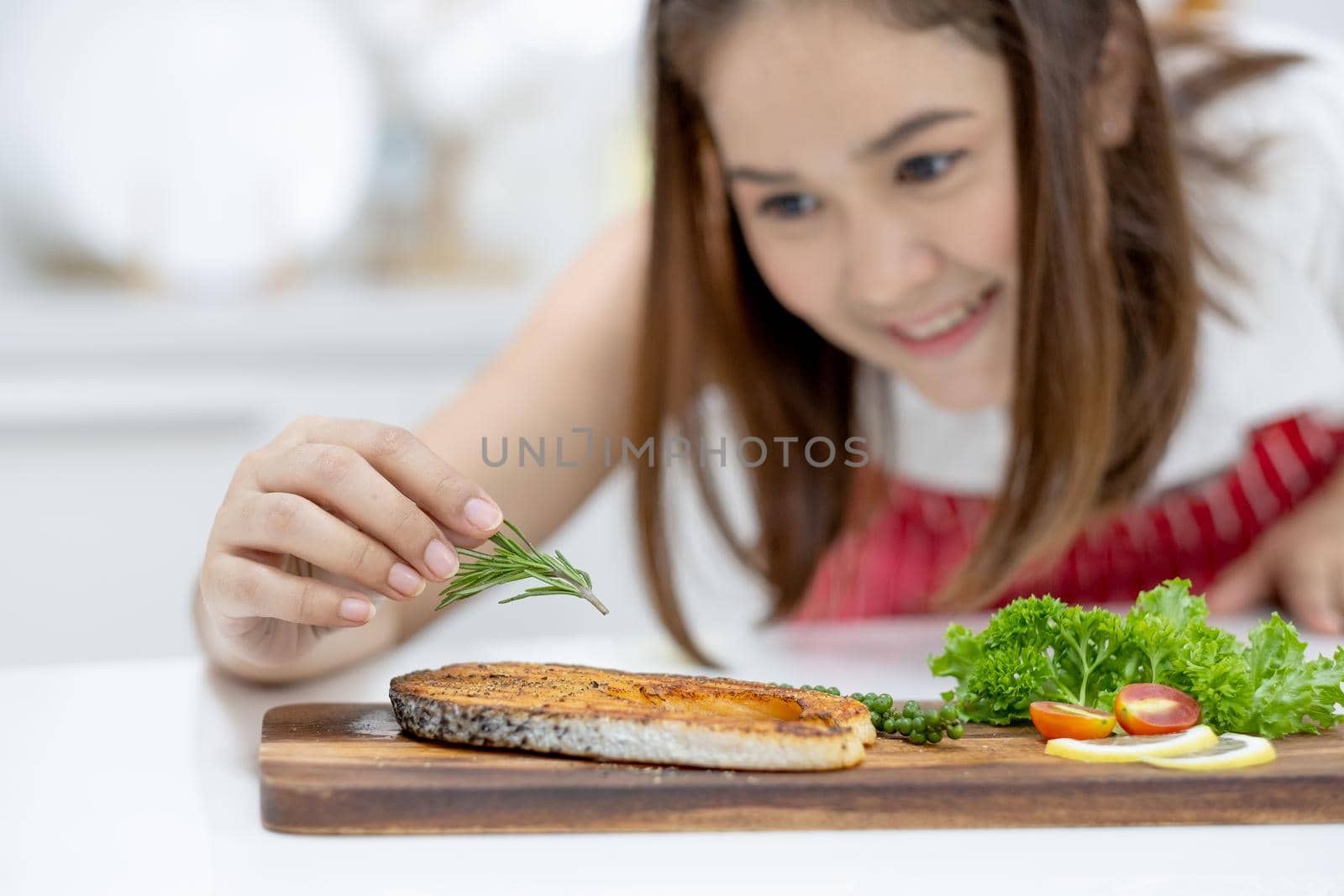 Asian beautiful woman with red apron put rosemary on slicing fish on plate in kitchen on table and her face show smiling of happy and proud emotion. Concept of happiness of cooking in their house.