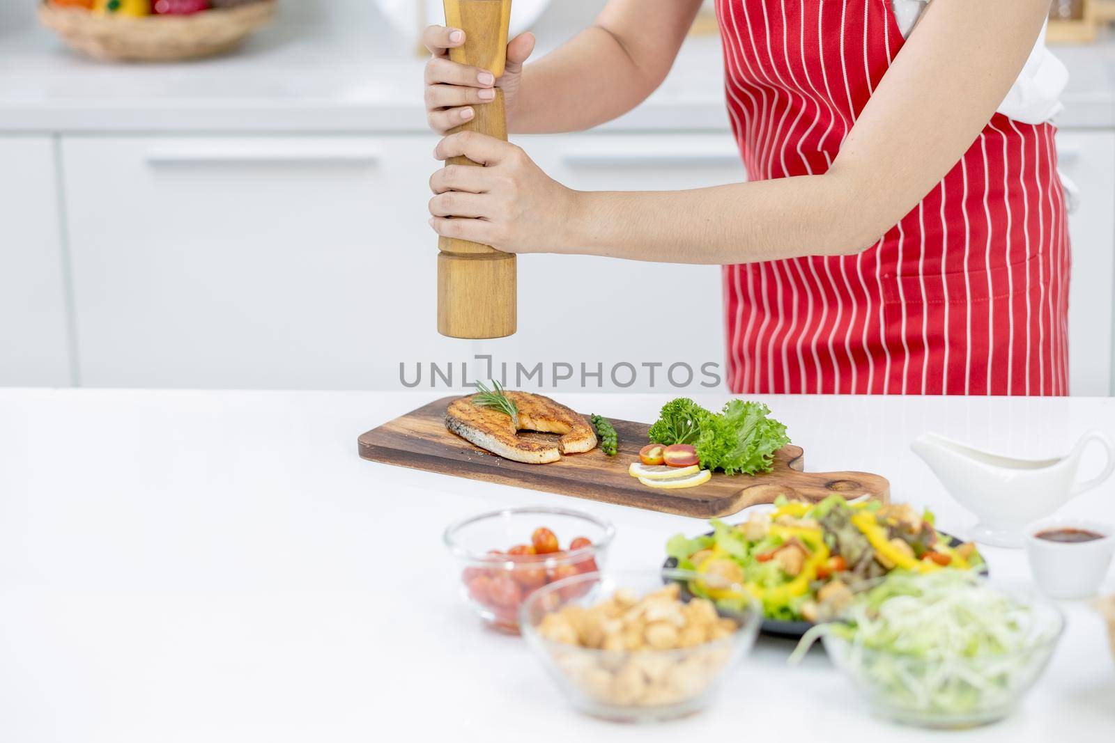 Hands of woman with red apron scatter spices over plate of slicing fish in kitchen with different type of ingredients and garnish on table. Concept of happiness of cooking in their house.