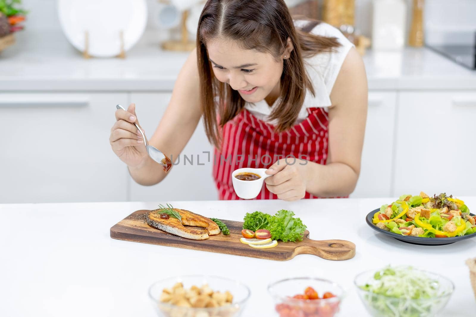 Asian beautiful woman with red apron drop tomato sauce on slicing fish on plate in kitchen with different types of bowl of ingredients on table. Concept of happiness of cooking in their house.