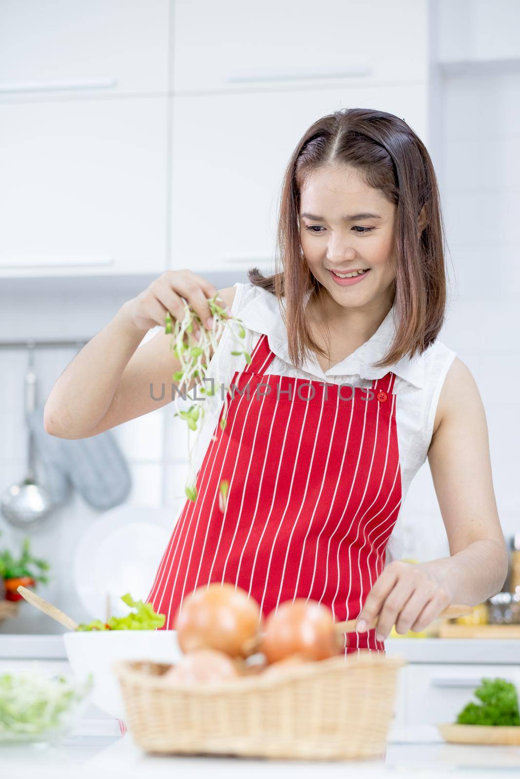 Asian beautiful woman with red apron scatter sprouts over bowl of vegetable salad in kitchen with different type of vegetable and garnish on table. Concept of happiness of cooking in their house.