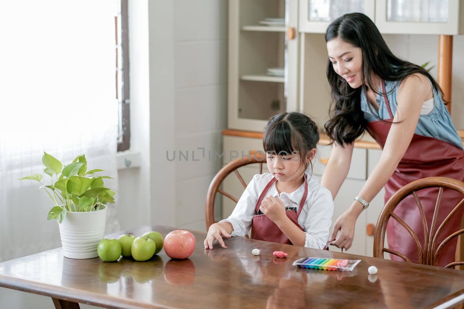 Little girl enjoy to play with play dough in kitchen on the table while her mother try to dress her with apron. Asian family with happiness concept.