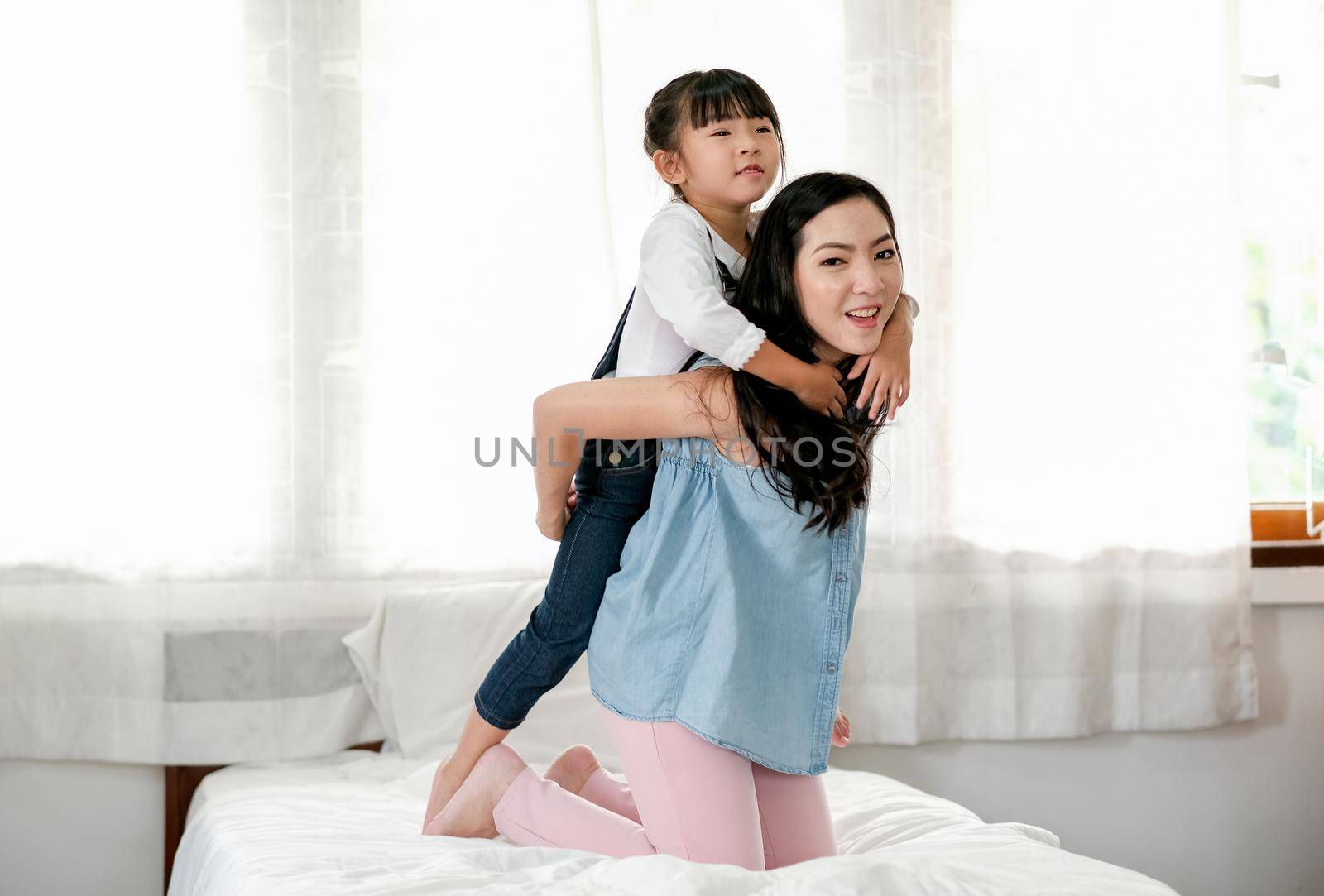 Asian mother and little girl have fun by girl hug on back of mother and stay on bed with happy and relax emotion.