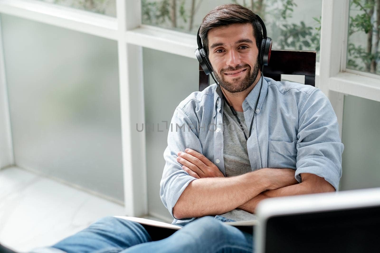 Caucasian man listen music by headphone and sit in room with glass windows with relax and happy expression also look at camera in room with day light.