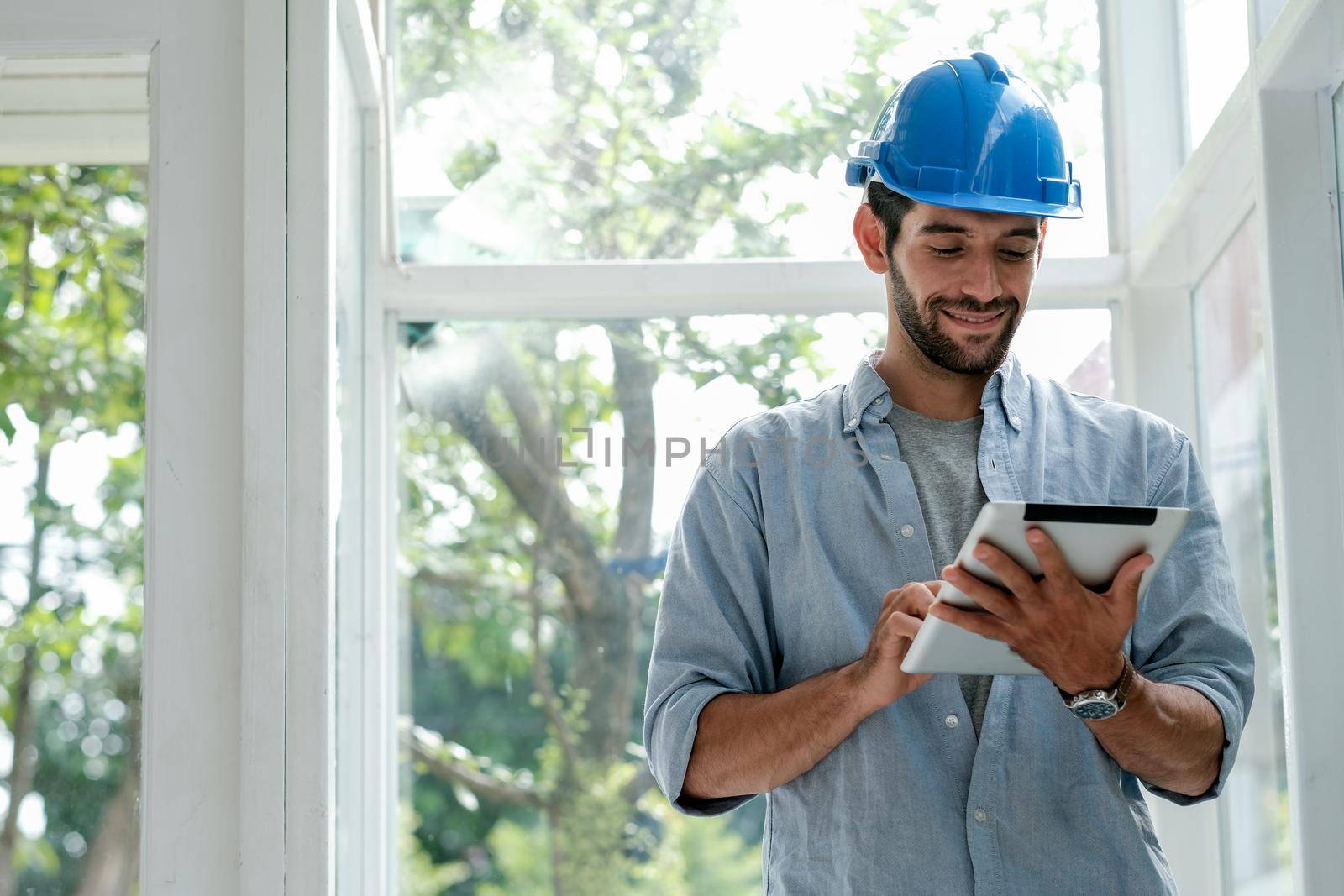 Caucasian engineer man with blue hardhat use tablet in office with glass window and he look happy with work.