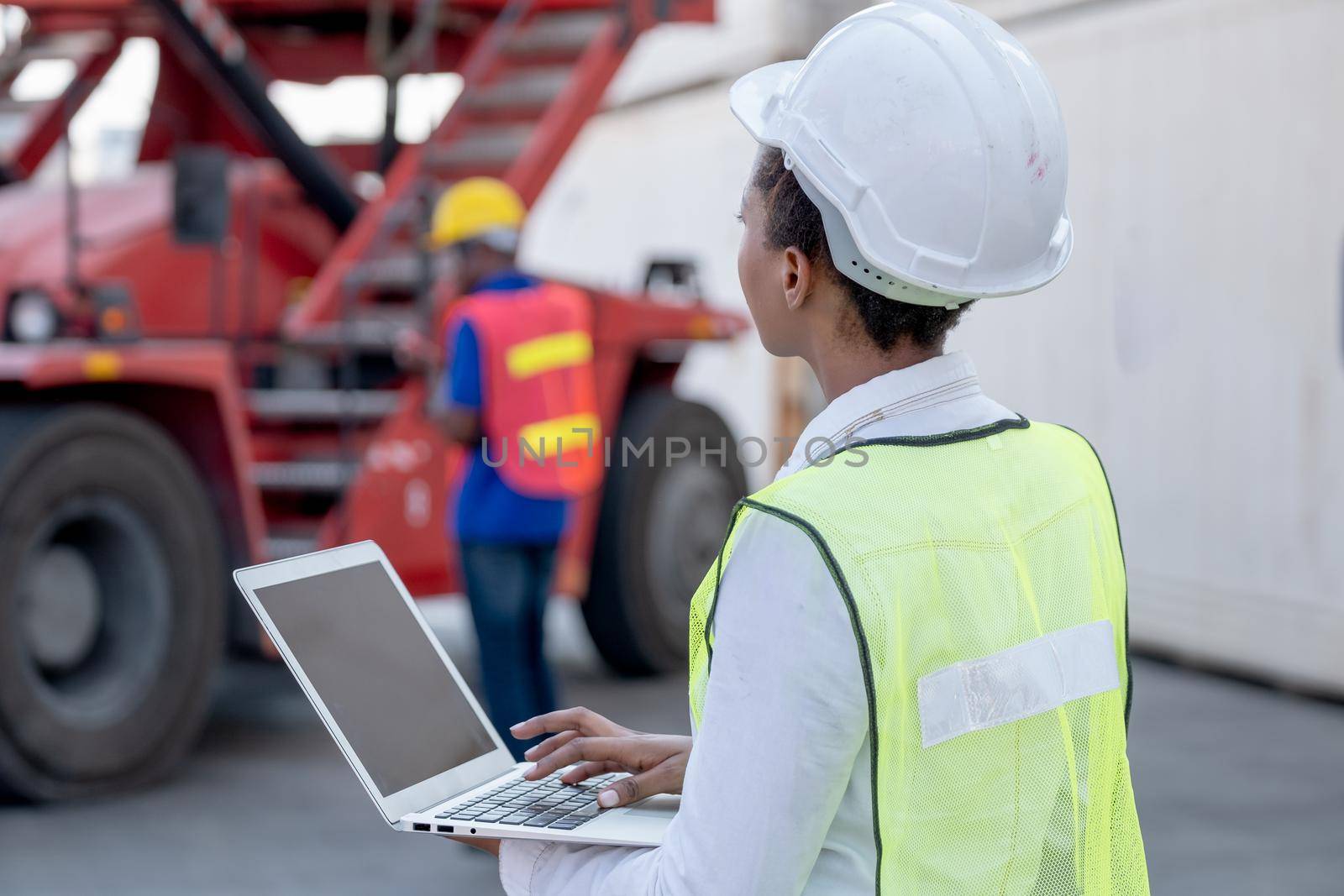 American African technician worker woman typing and look to truck while her co-worker work with construction vehicle. Concept of good system and manager support for better industrial business.