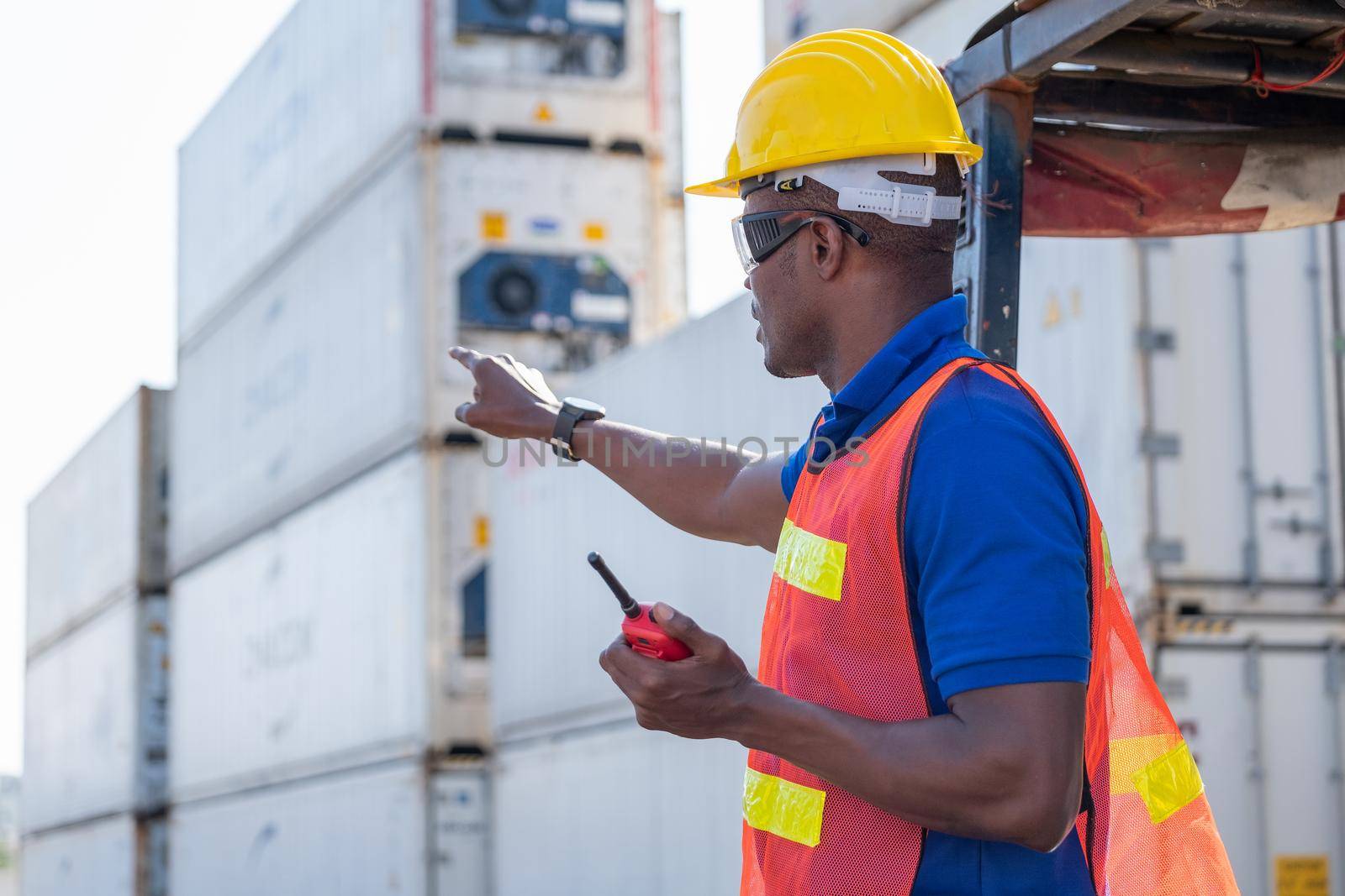 African American foreman or cargo container worker hold walkie talkie and point to front direction look like communicate with his team to work in workplace.