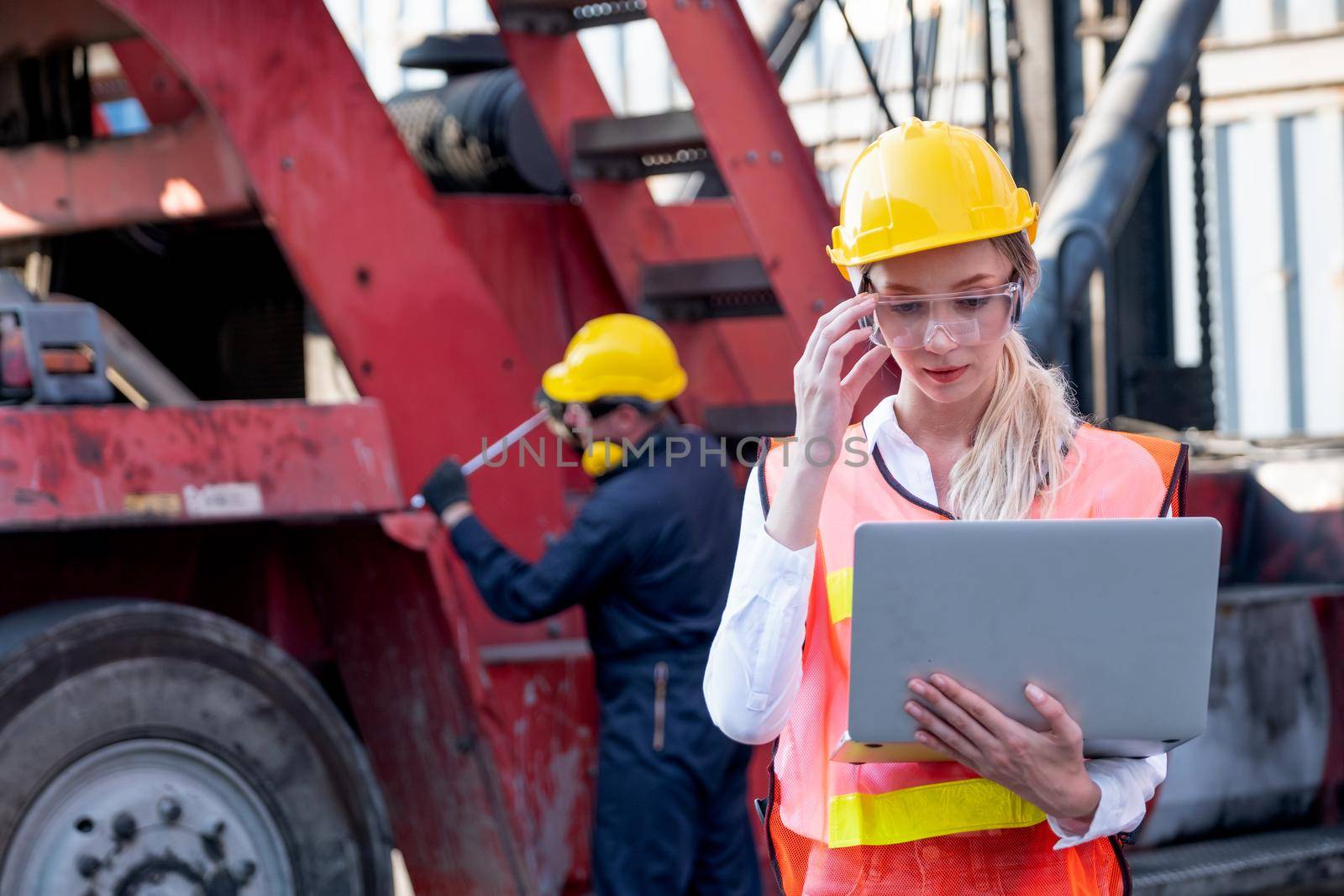 Pretty foreman woman or worker stand with touch her safety eye glasses and holding laptop for working in front of her co-worker fix the problem of vehicle in background of cargo container workplace. by nrradmin