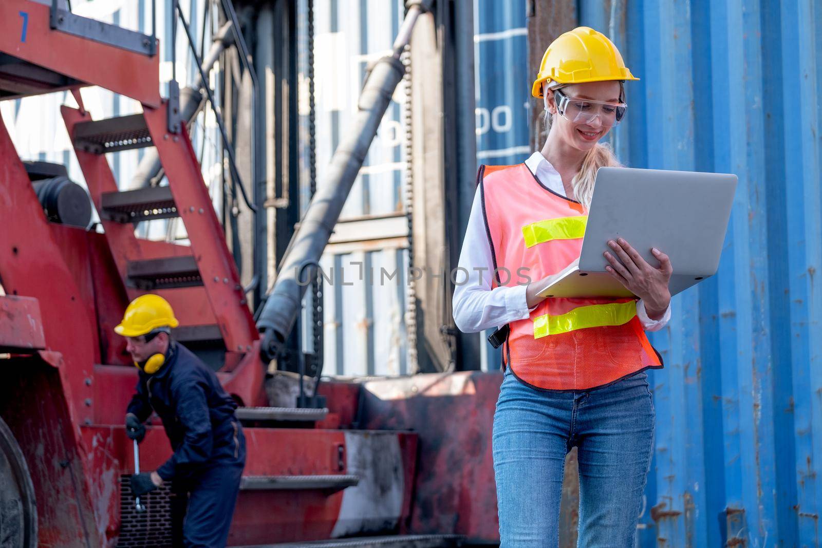 Pretty foreman woman stand with holding laptop for working in front of her co-worker fix the problem of vehicle in background of cargo container workplace.