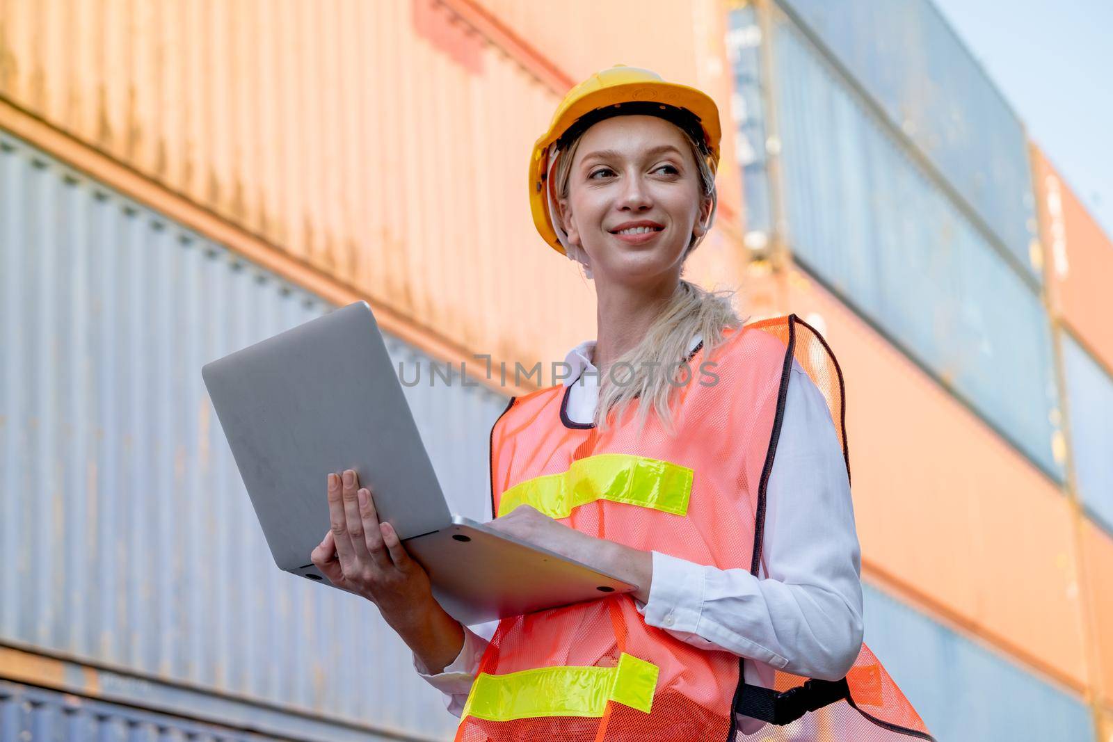 Beautiful foreman woman hold laptop and smile during working in cargo container workplace with background container stack with multi-color. Industrial system support employee and staff work concept.