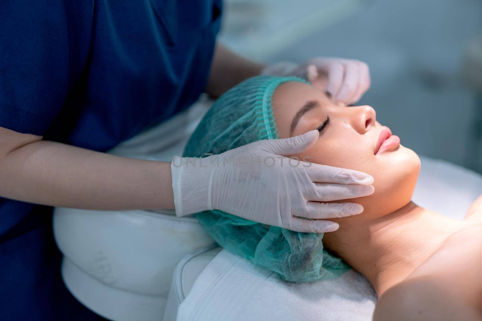 Beauty clinic hands touch on face of customer woman who lie on bed with light during process of treatment.