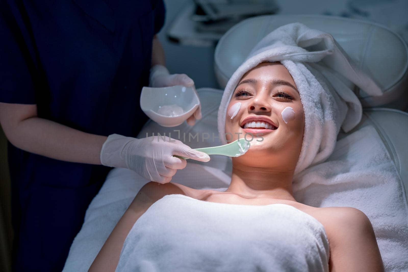 Wide shot hands hold paddle apply beauty cream on face of beautiful woman during process in beauty clinic or salon.