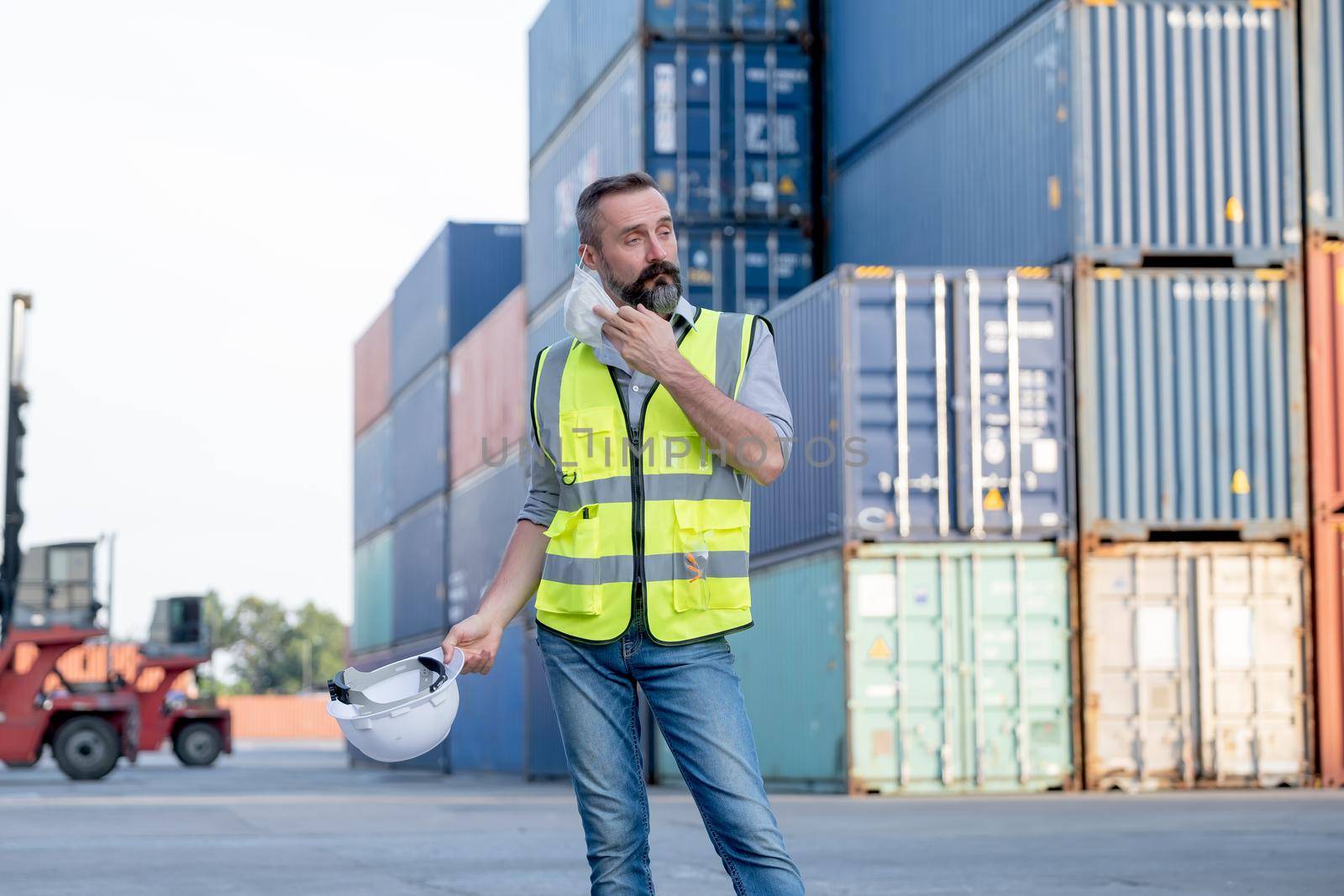 Factory foreman of cargo container worker man take of mask and hardhat after finish work. Concept of new normal lifestyle in industrial business during pandemic of Covid-19 coronavirus in city.