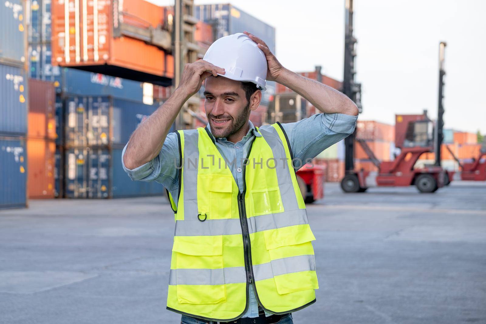 Foreman engineer or cargo container worker hold hardhat and stand in front of cranes and stack of shipping. Logistic shipyard management system and safety concept.