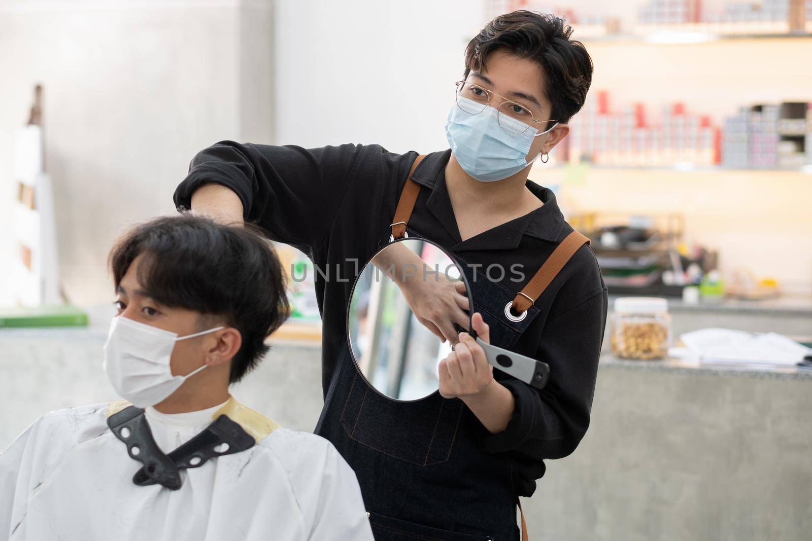 Beauty salon Asian barber man with hygiene mask use mirror to show hairstyle to customer in the shop. by nrradmin